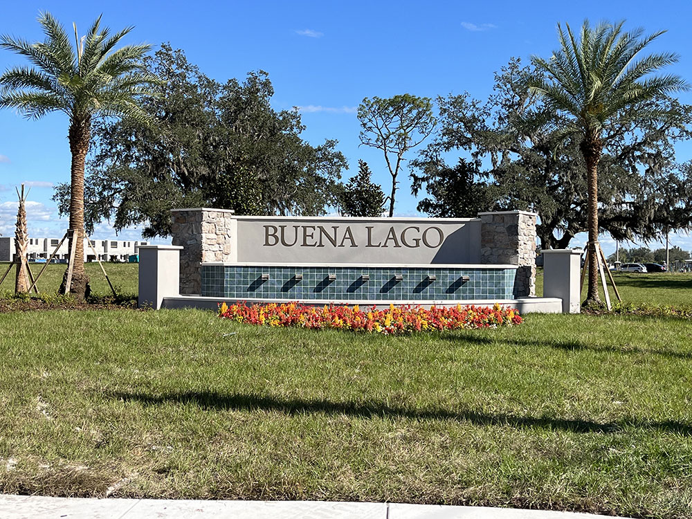 Large concrete sign with a pillar of stone veneer on each side. Sign is surrounded by grass and 1 palm tree on each side.