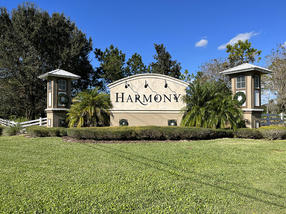 Large sign with "Harmony" in center. 1 pillar on each side with small palm trees and bushes around sign.