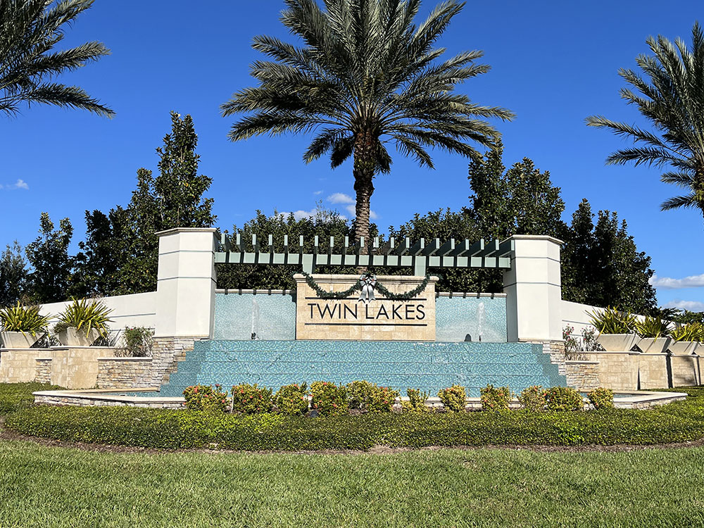 Sign at entrance of Twin Lakes Community in St. Cloud at Hickory Tree Road. The area is home to many new communities drawing St Cloud Florida Real Estate buyers. Palm trees are towering over the sign and 2 small fountains are shooting water up on each side of "Twin Lakes".The fountains drain forward to create a shelved water fall.