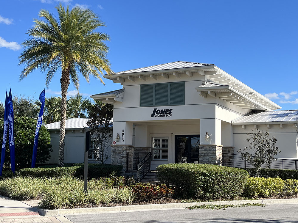 Brand new building with pillars of stone halfway up. Text on building reads "Jones Homes USA" and the address "4910. A tall green palm tree is in front of the building and a varierty of shrubs and plants surround the entrance.